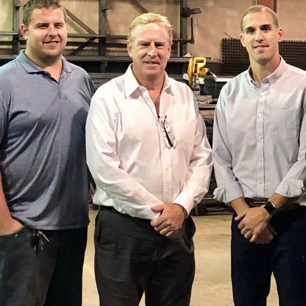 Three men standing in a warehouse with one man holding his hands on the other mans shoulder.